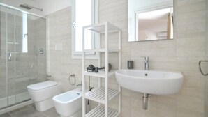 Et moderne badeværelse med et glasafskærmet brusebad, en hvid konsolvask og en hvid kommode med hylder.