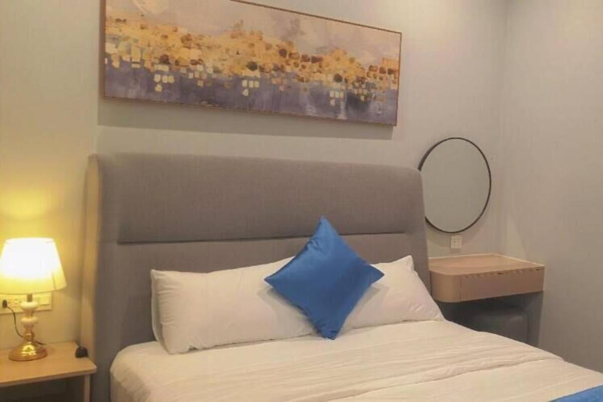 A hotel room with a bed, a bedside table with a lamp, a round mirror, and a painting on the wall.