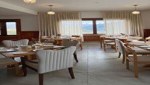 A dining area with multiple tables and chairs, large windows with curtains, and hanging light fixtures. - The Zurfi's Hotel (Kargil)