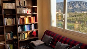 Eine gemütliche Leseecke mit Bücherregal, einer rot-schwarzen Couch und Blick auf Berge durch ein Fenster.
