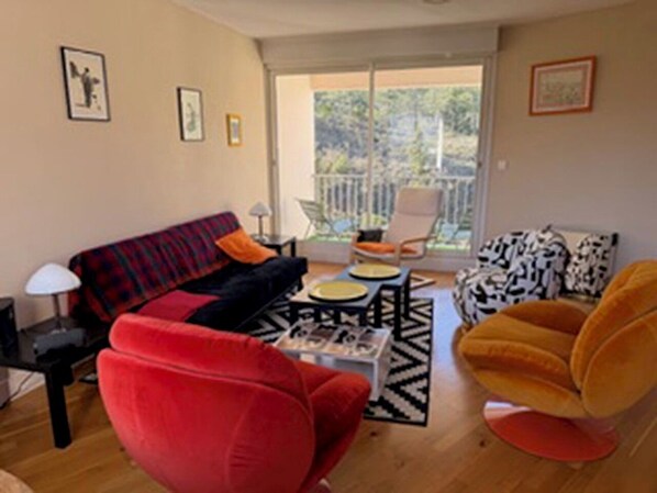 Una sala de estar con un sofá, un sillón rojo, un sillón amarillo y una mesa de centro. Hay un balcón con una silla y una mesa.