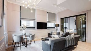A modern living room with a grey sofa, a dining table with chairs, a flat-screen TV, and large windows with blinds. - Central 2-Bed, Walk to Chinatown & Theatres (Greater Manchester)