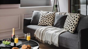 A modern living room with a grey sofa, polka dot cushions, a blanket, and a small table with croissants, grapes, champagne, and orange juice. - Central 2-Bed, Walk to Chinatown & Theatres (Greater Manchester)