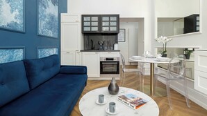 A modern living room with a blue sofa, a round coffee table with a vase and a magazine, and a kitchen with white cabinets and a black range hood. - Climatic Studio | Close to Market Square and Wawel Castle (Kraków)