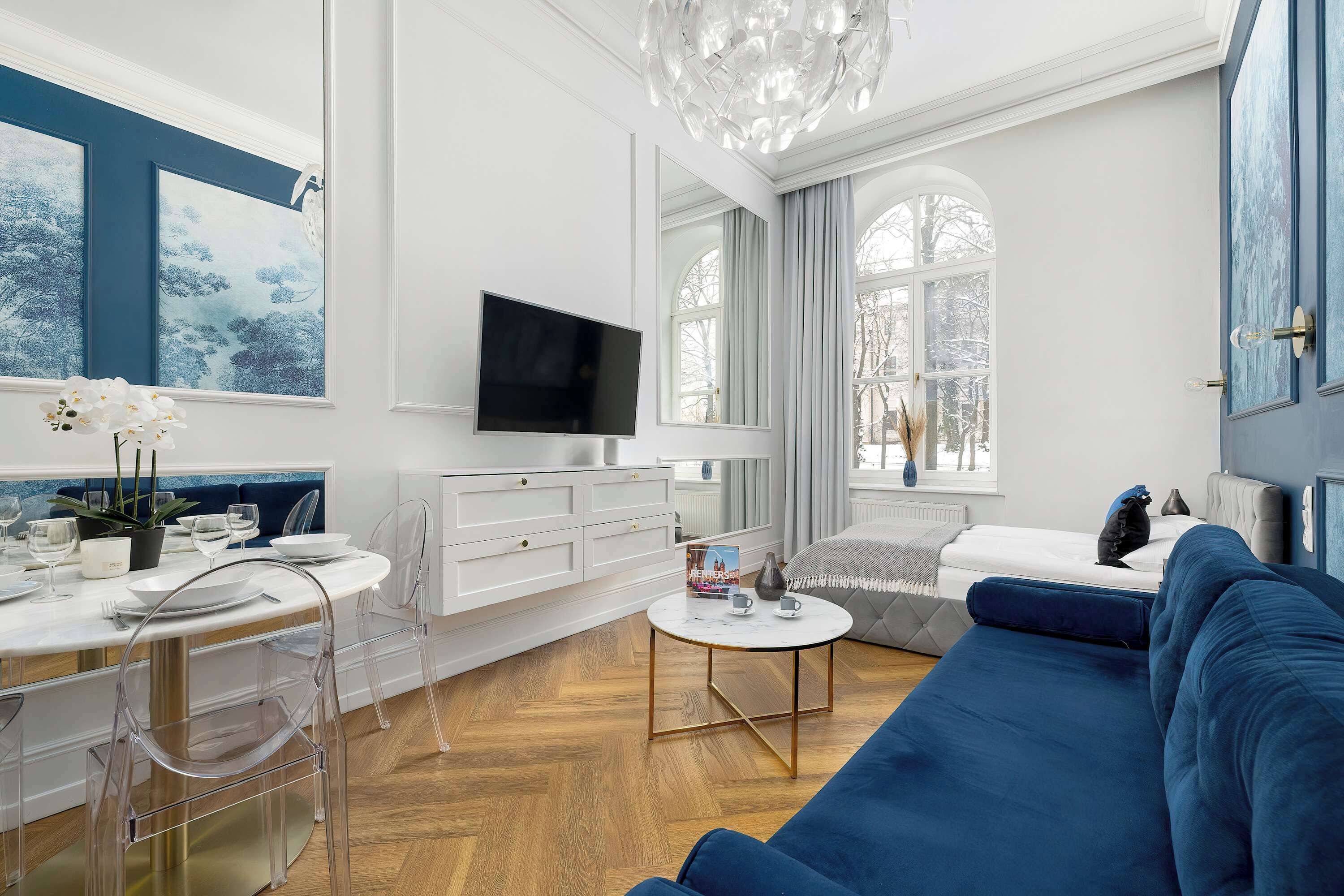 A modern living room with a blue sofa, a white dining area, and a TV on the wall.