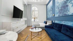 A modern living room with a white marble coffee table, a blue sofa, a flat-screen TV, and a large window with a view of trees. - Climatic Studio | Close to Market Square and Wawel Castle (Kraków)