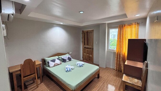 Quarto pequeno e limpo com uma cama de solteiro, uma escrivaninha de madeira, uma cadeira, uma televisão e uma janela com cortinas.