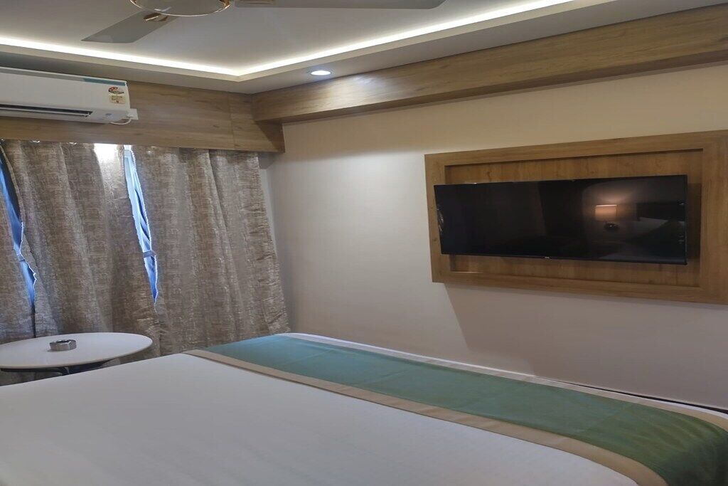 A hotel room with a bed, a TV mounted on the wall, an air conditioning unit, a small round table, and curtains.