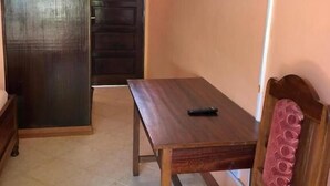 A small room with a wooden table, a chair with a patterned cushion, a TV mounted on the wall, and a door leading to another room.