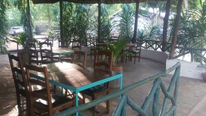 An outdoor patio area with wooden tables and chairs, surrounded by potted plants and trees.