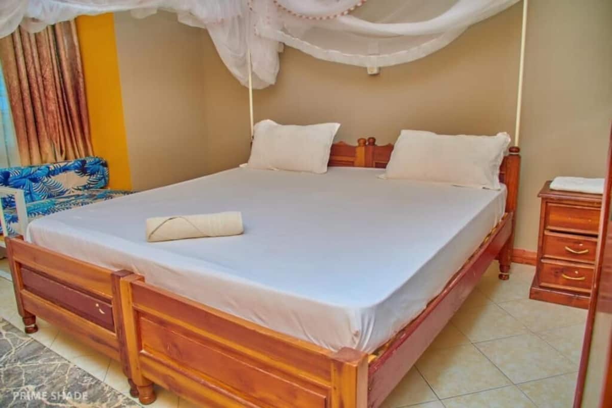 Una cama bien hecha con cubrecama y almohadas blancas, cabecera de madera y mesita de noche con cajones.