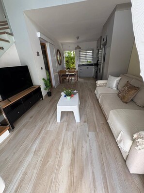 Ein modernes Wohnzimmer mit Fernseher, einem weißen Couchtisch, einem beige gefärbten Sofa und einer Küche im Hintergrund.