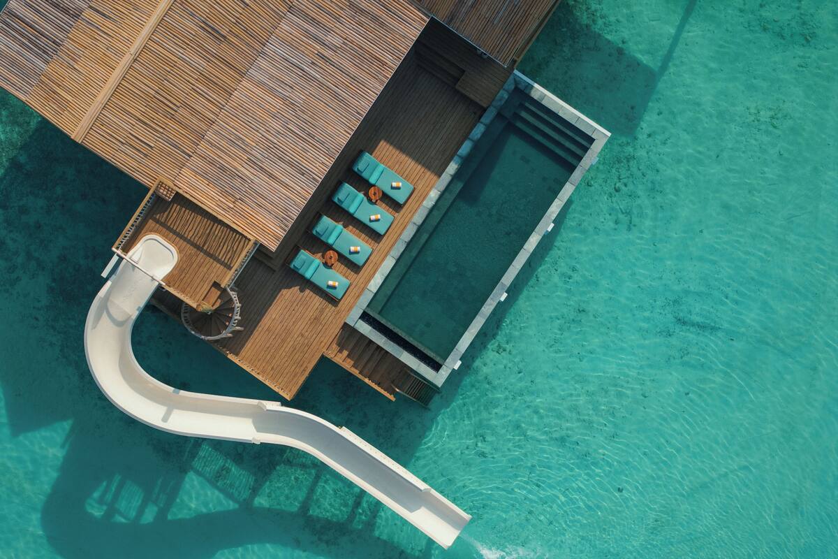 Vista aérea de un complejo turístico con piscina, zona de descanso y tobogán que desemboca en el agua.