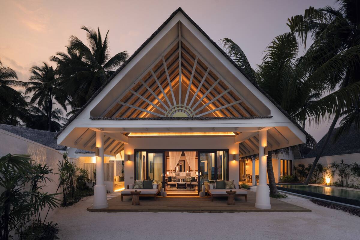Un resort con un espacio de estar techado al aire libre, piscina y palmeras.