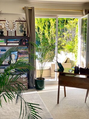 Ein Zimmer mit einem großen Schiebeglasfenster, das in einen Garten führt, einem Bücherregal voller Bücher, einer Topfpflanze und einem Esstisch.