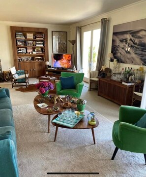 Ein Wohnzimmer mit einer Couch, einem grünen Sessel, einem Couchtisch, einem Bücherregal, einem Fernseher und einem Bild an der Wand.