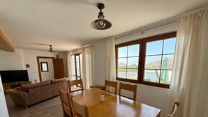 A dining area with a wooden table and chairs, a sofa, a television, and a view of a mountain through large windows. - Casa Joanna Lanzarote (Nazaret)