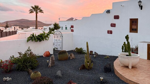 A rooftop garden with cacti and plants, a white wall with a gate, and a view of distant buildings and hills. - Casa Joanna Lanzarote (Nazaret)