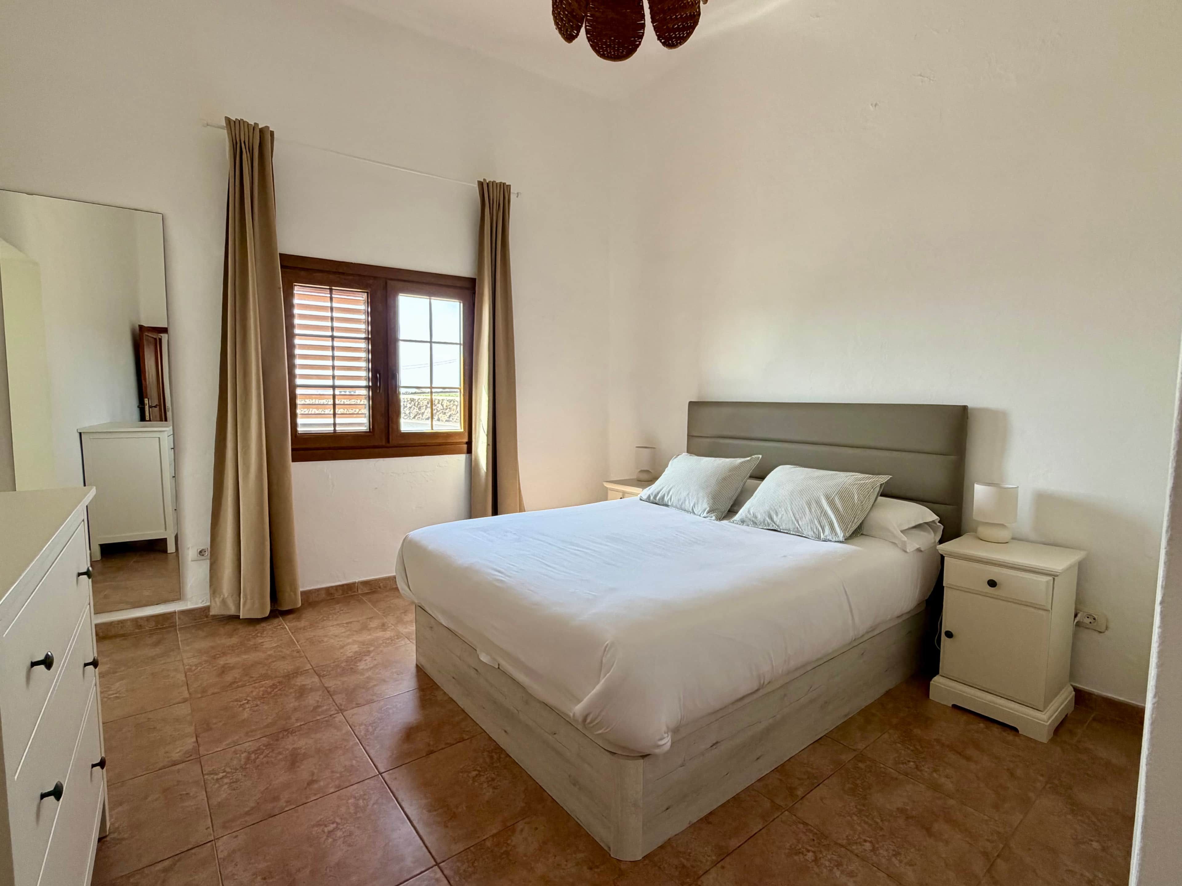 A bedroom with a bed, nightstands, a dresser, a window with curtains, and a ceiling fan.