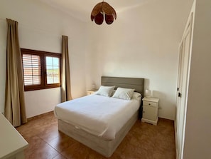 A bedroom with a bed, bedside tables, a window with curtains, and a hanging light fixture. - Villa Ann Lanzarote (Nazaret)