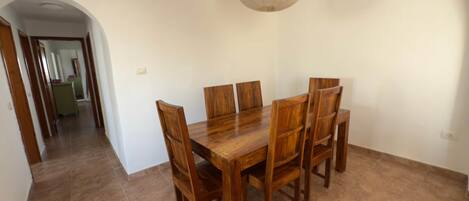 A dining area with a wooden table and chairs, a hanging light fixture, and a hallway leading to another room.