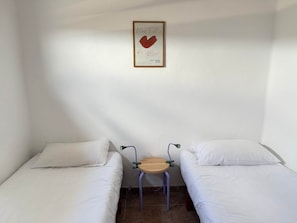 Quarto pequeno com duas camas de solteiro, uma mesinha e um letreiro fixado na parede.