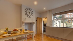 A modern kitchen with a wooden dining table, a large wall clock, and a view of the outside through a window. - Beecroft Cottage - sleeps 4 guests  in 2 bedrooms (Cromer)