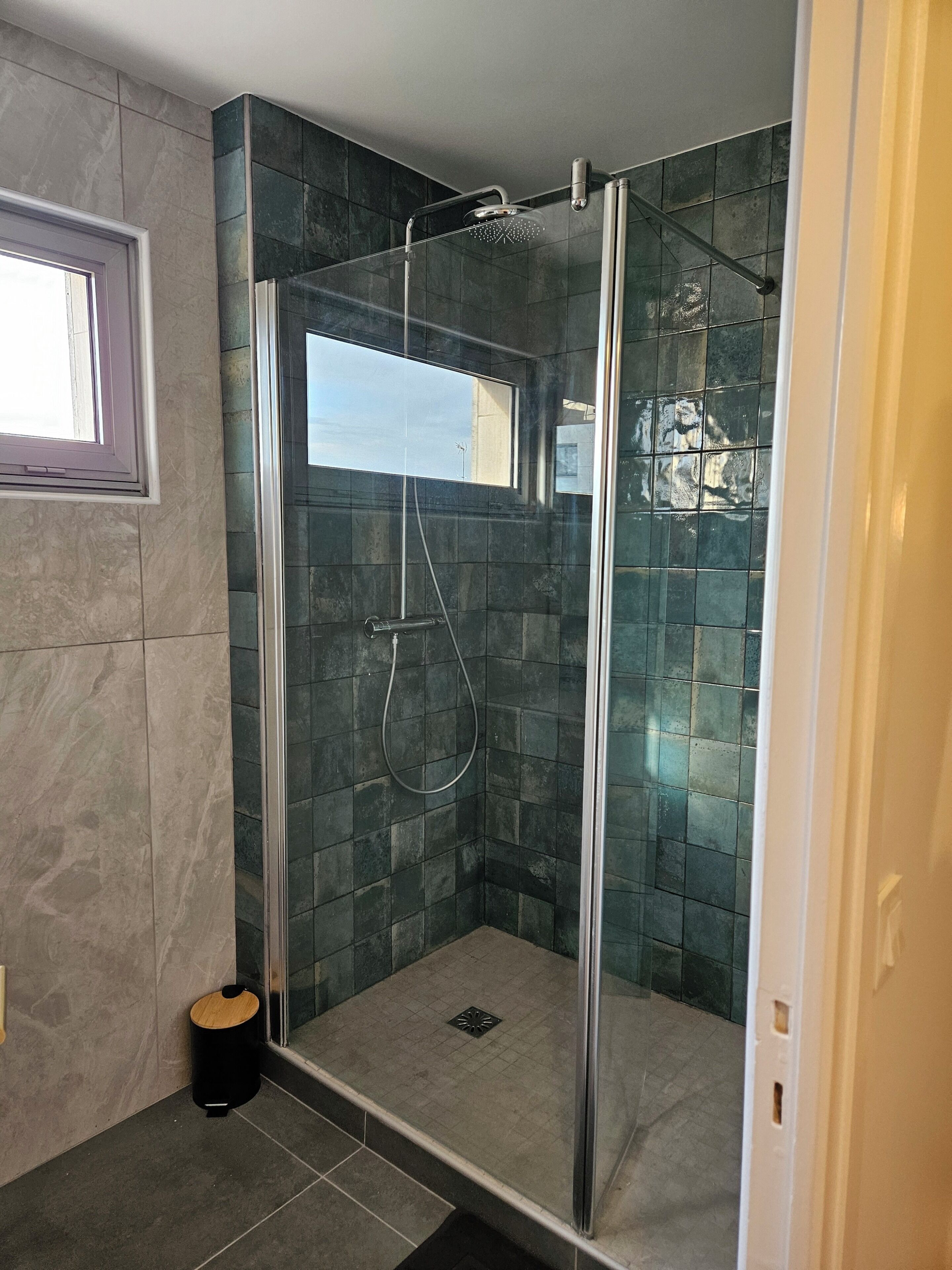 A glass-enclosed shower with a window, tiled walls, and a small trash bin.