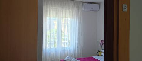 A bedroom with a bed, a window with curtains, an air conditioning unit, and a door.
