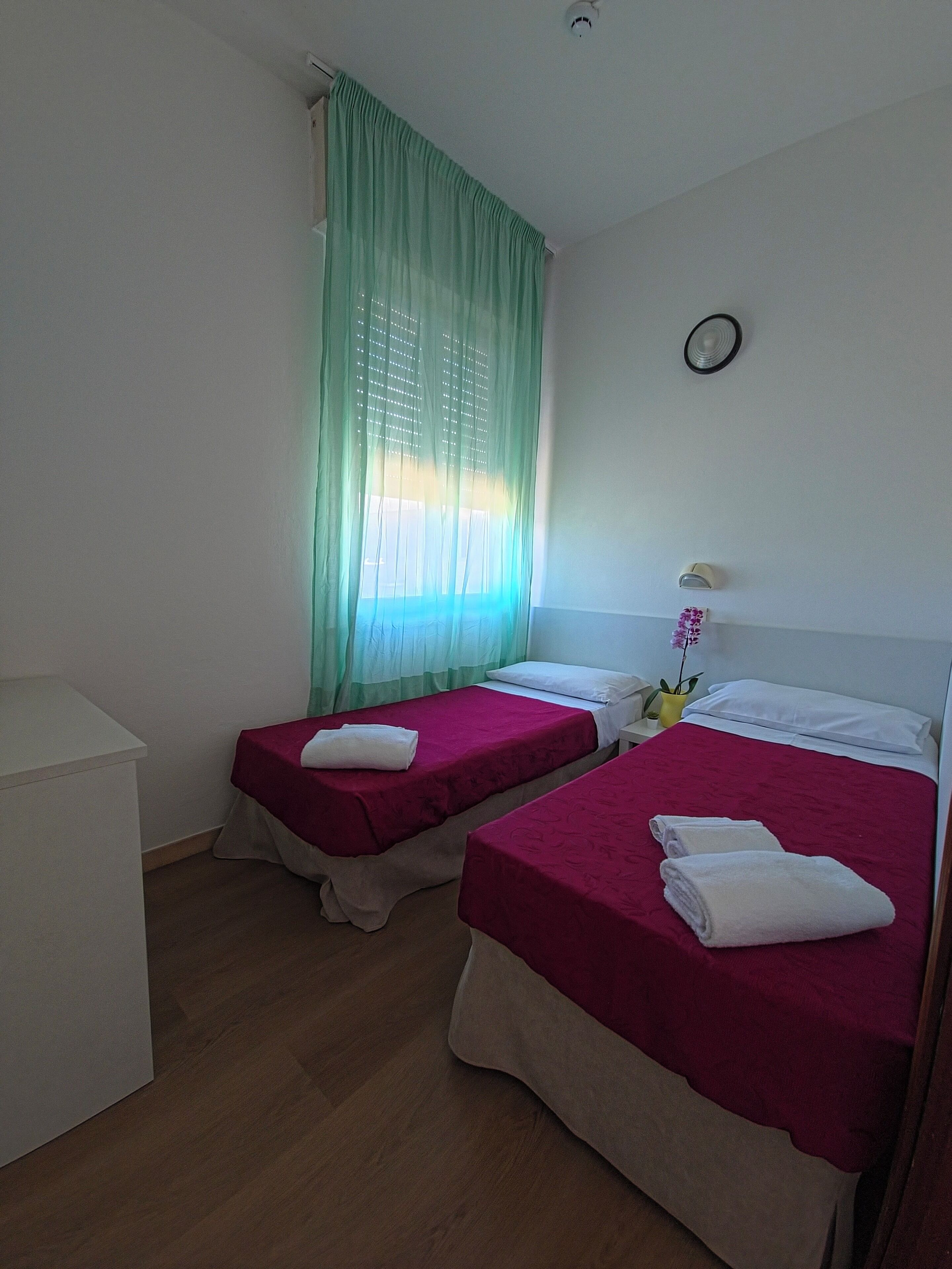 Quarto com duas camas, uma escrivaninha, janela com cortinas leves, relógio na parede e um pequeno vaso com flores.