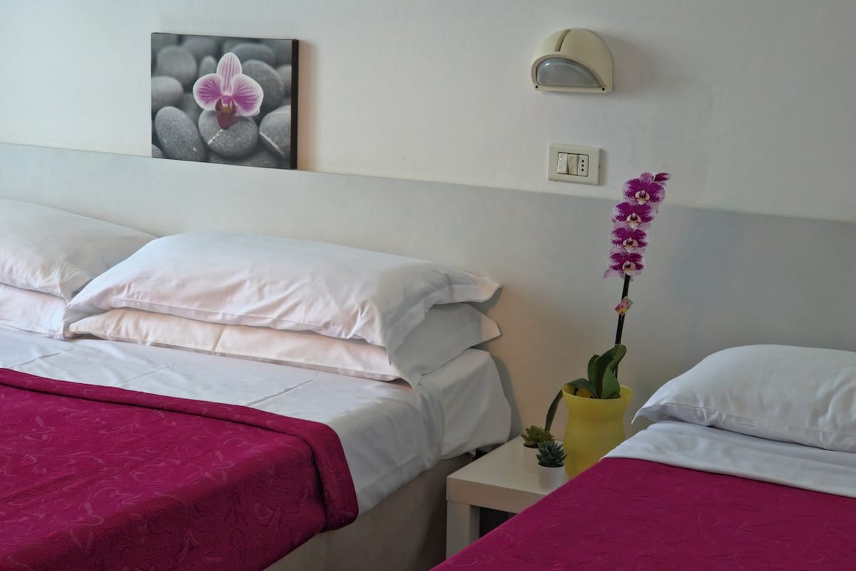 Habitación de hotel con dos camas, una mesita de noche con lámpara, un interruptor en la pared y un cuadro con una orquídea.