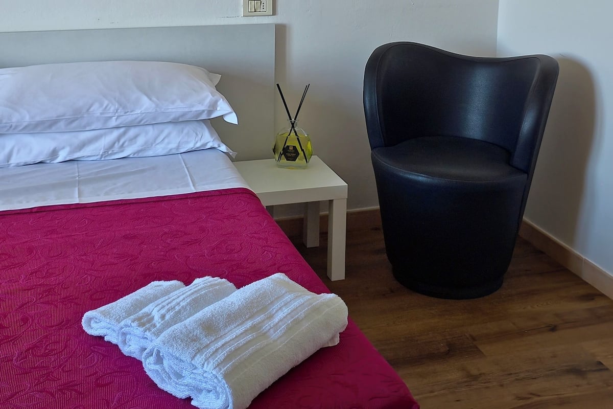 Una habitación con una cama, una silla, una mesita de noche, una lámpara y una luz empotrada en la pared.
