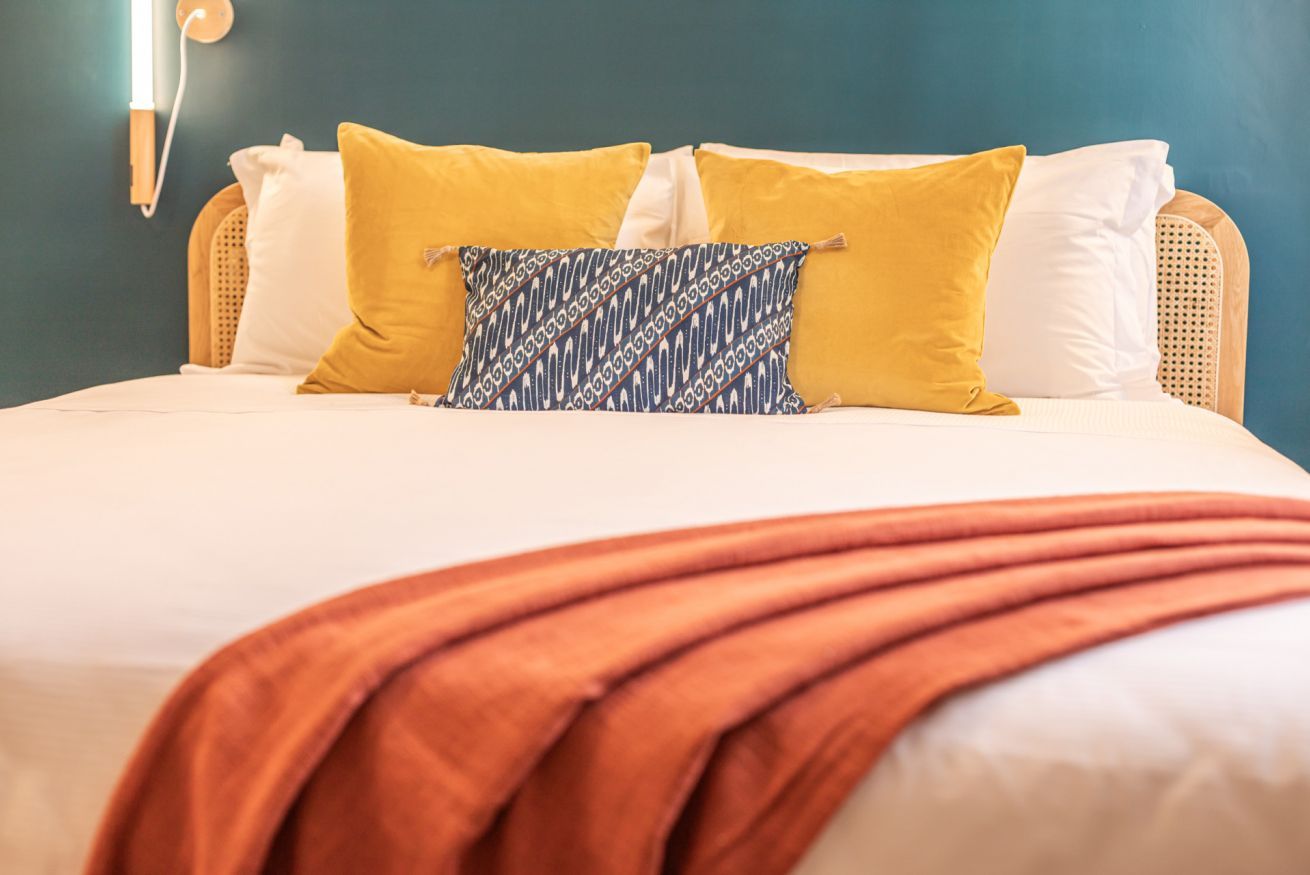 A bed with a white mattress, a brown blanket, and multiple pillows in yellow and patterned designs.