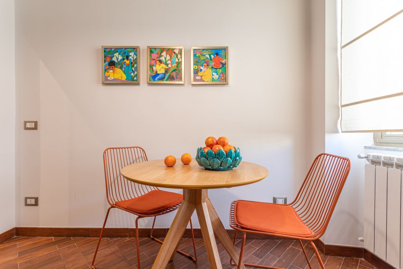 Un comedor con una mesa redonda de madera, dos sillas naranjas y un cuenco de naranjas. Tres cuadros coloridos cuelgan de la pared sobre la mesa.