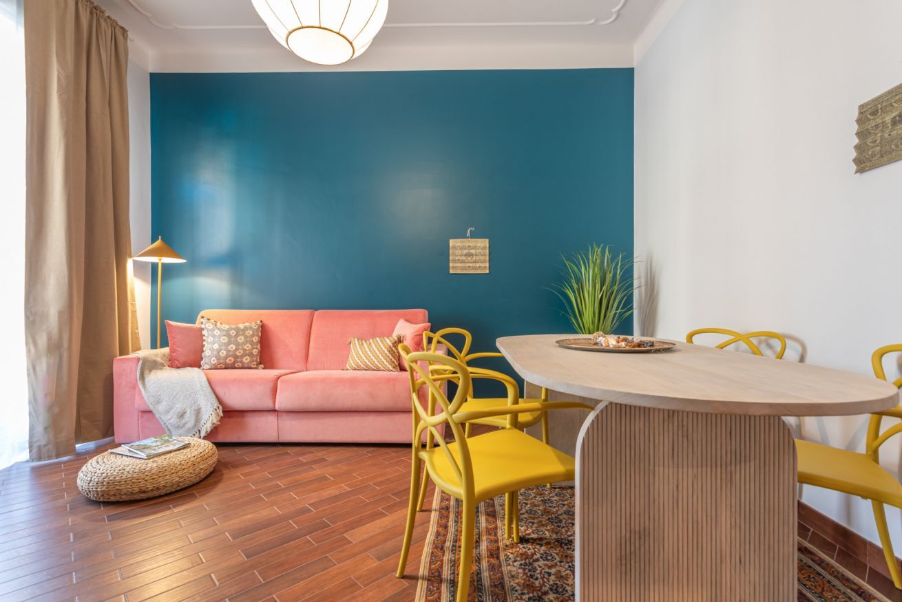 Ein Wohnzimmer mit blauer Akzentwand, einem rosafarbenen Sofa, gelben Sesseln und einem Holzesstisch.