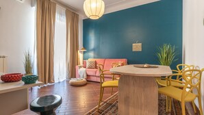 Ein modernes Wohnzimmer mit blauer Akzentwand, einem runden Holztisch, gelben Stühlen, einem rosafarbenen Sofa und einem großen Fenster mit beigen Vorhängen.