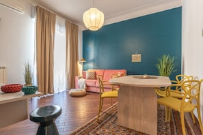 Ein modernes Wohnzimmer mit blauer Akzentwand, einem runden Holztisch, gelben Stühlen, einem rosafarbenen Sofa und einem großen Fenster mit beigen Vorhängen.