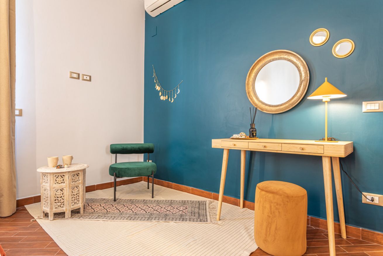 A room with a blue accent wall, a round mirror, a small table with a lamp, a green chair, and a small stool.