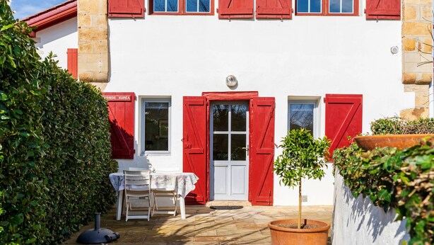 Un bâtiment avec des volets rouges, une porte blanche et une petite table avec des chaises à l’extérieur.