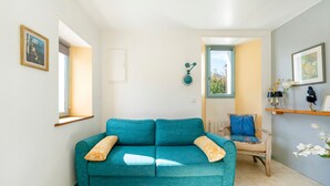 Un canapé de couleur turquoise agrémenté de coussins à motifs, une chaise en bois avec un coussin bleu, une petite table sur laquelle est posé un vase, et une fenêtre donnant sur un paysage verdoyant.