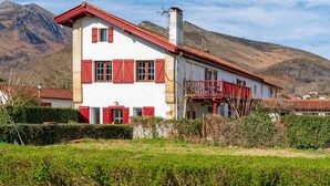 Une maison à deux étages, avec un toit rouge et des murs blancs, entourée de verdure et de montagnes.