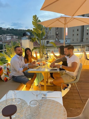 A group of people dining outdoors on a wooden deck with a table set for a meal, surrounded by large umbrellas and plants. - suite la pennichella (Agropoli)