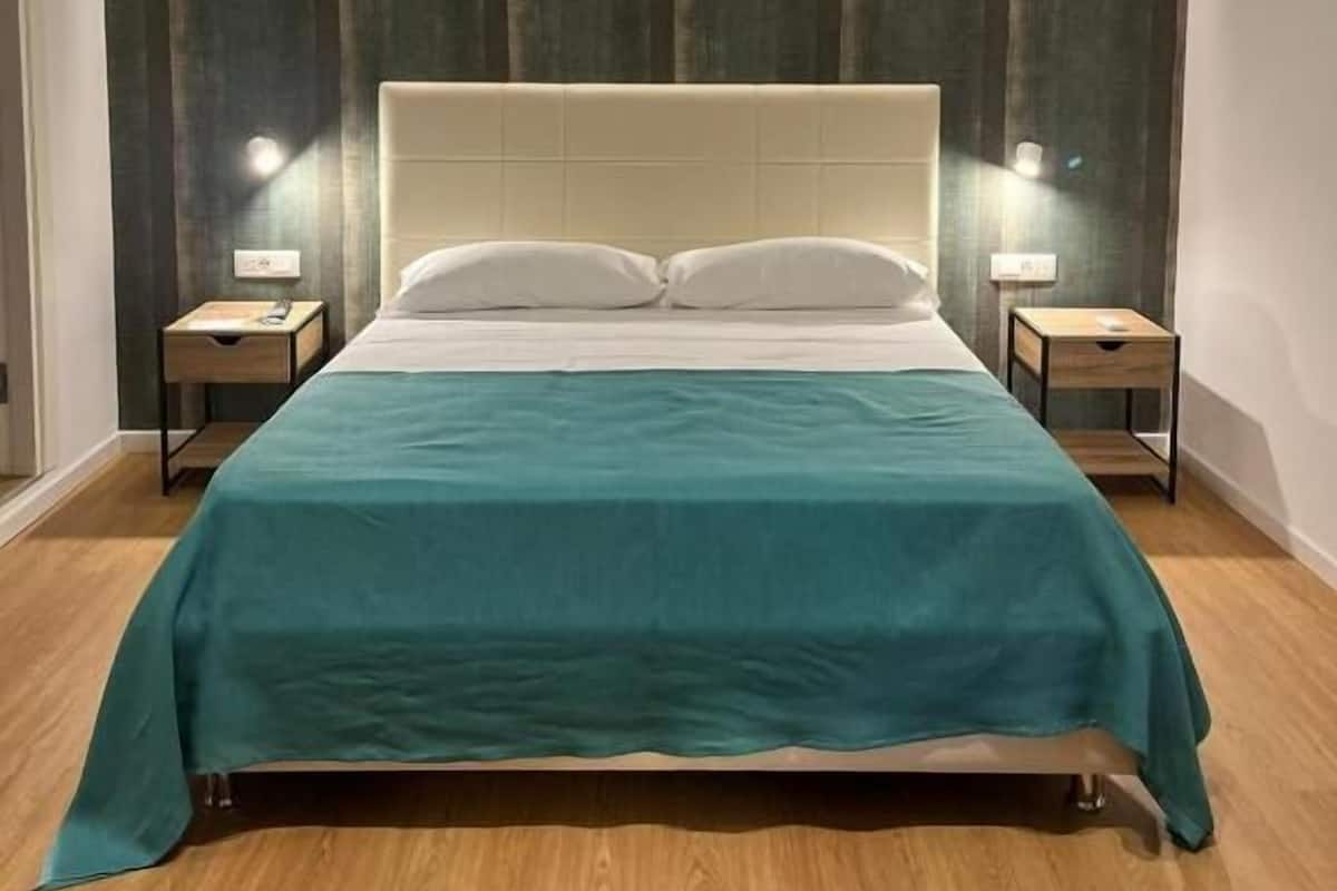 A hotel room with a bed, two bedside tables, a nightstand, a wall-mounted light, and a curtain.