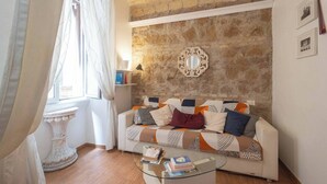A cozy living room with a stone wall, a white sofa with patterned cushions, a glass coffee table, and a white pedestal table. - Colosseum: Entire Guest House (2BR en-suite/2BA) (Roma)