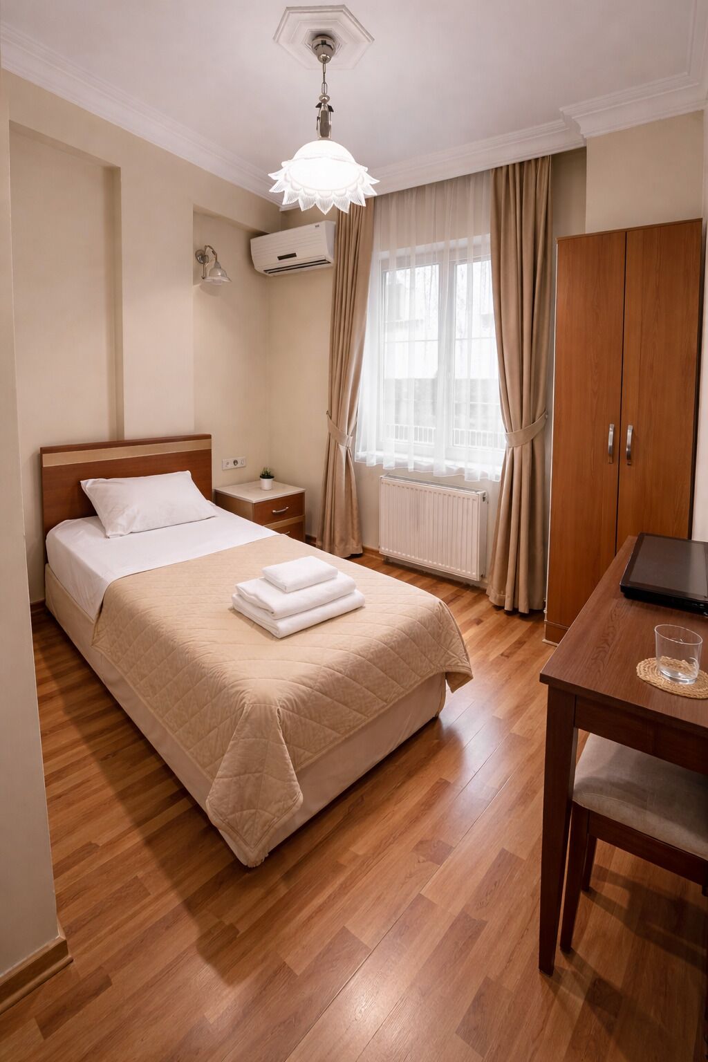 Habitación de hotel con cama, mesita de noche, lámpara, ventana con cortinas y escritorio con computadora portátil.