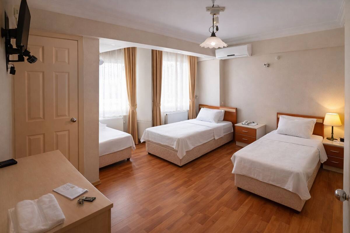 A hotel room with two single beds, a desk with a chair, a bedside table with a lamp, and a ceiling-mounted air conditioning unit.
