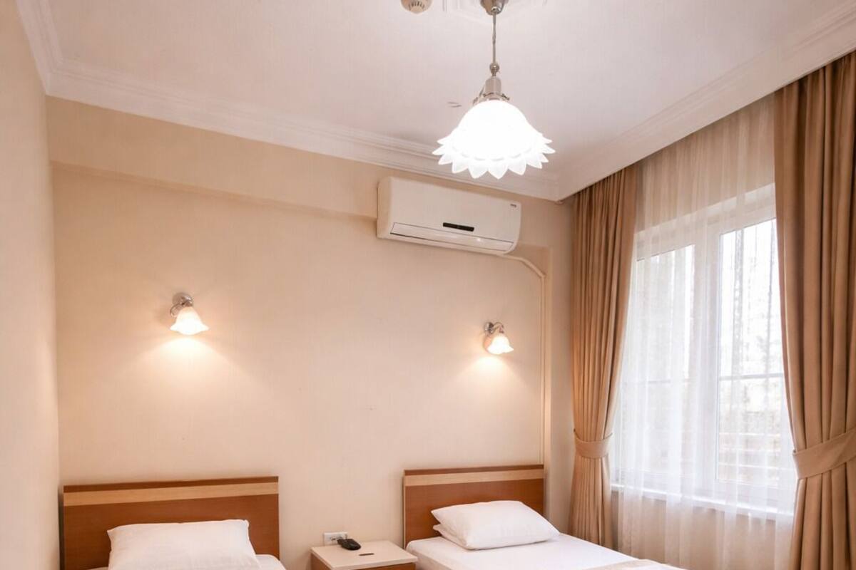 A hotel room with two beds, a nightstand, an air conditioning unit, and a window with curtains.