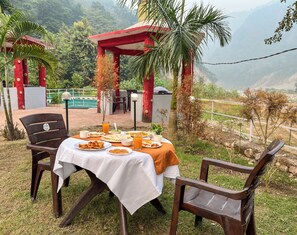 Un espace de restauration en plein air avec une table dressée pour deux personnes, entourée de chaises, offrant une vue sur les montagnes et donnant sur une construction au toit rouge.