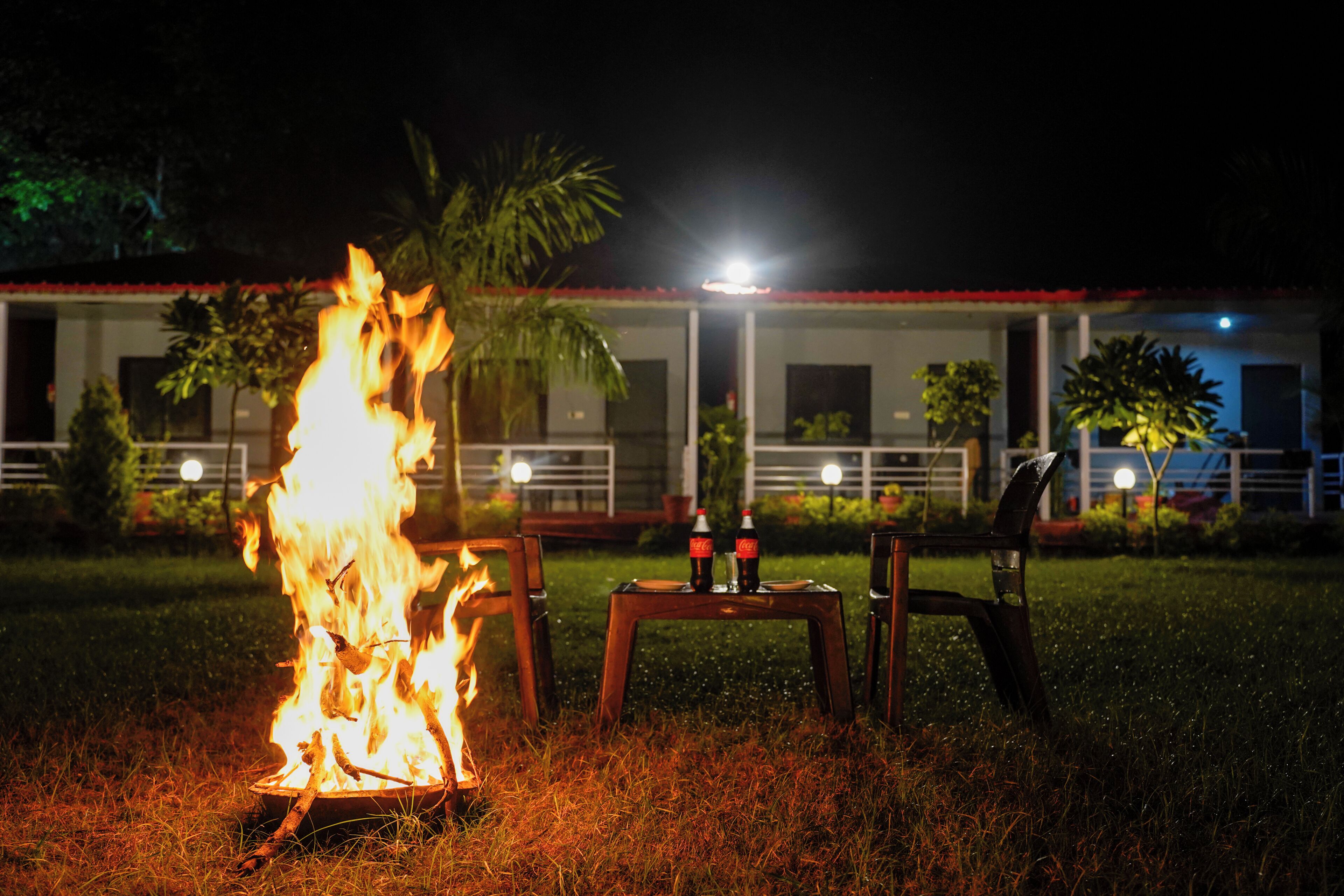 A nighttime outdoor setting with a fire, two chairs, and a table with bottles.