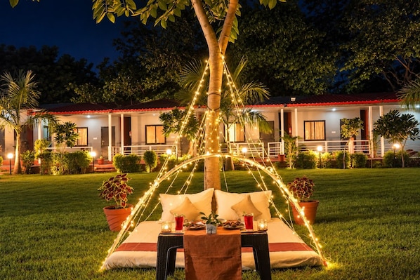 An outdoor dining area with string lights, a table set for two, and a building in the background. - Ekantam Riverside Resort (Rishikesh)